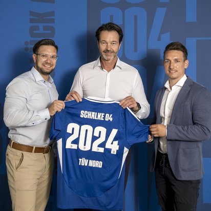 Drei Männer in Business-Kleidung stehen vor einer blauen Wand mit Schriftzügen und dem Logo von Schalke 04. Sie halten gemeinsam ein blaues Fußballtrikot mit der Aufschrift „Schalke 04“, der Jahreszahl „2024“ und dem TÜV NORD-Logo. Die Männer lächeln in die Kamera, während sie das Trikot präsentieren.