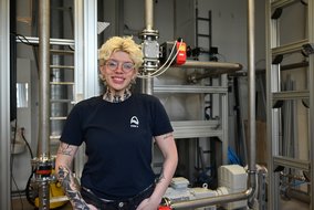 Liliana Quintero Zambrano lächelt in einem dunklen T-Shirt mit Logo vor einer technischen Anlage.