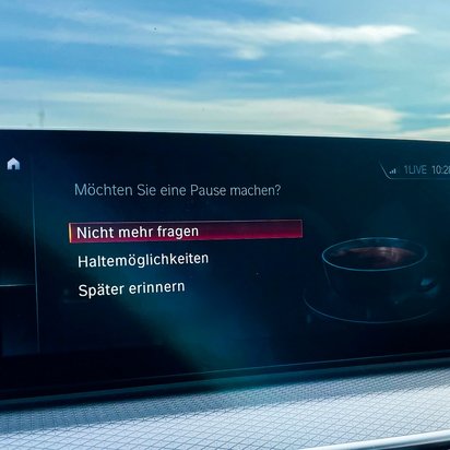 Autodisplay mit einer Sicherheitswarnung zur Fahrerermüdung. Der Bildschirm zeigt die Frage „Möchten Sie eine Pause machen?“ mit drei Auswahlmöglichkeiten: „Nicht mehr fragen“, „Haltemöglichkeiten“ und „Später erinnern“. Im Hintergrund ist eine stilisierte Kaffeetasse zu sehen. Das Auto befindet sich in Bewegung, mit einer Landstraße und blauem Himmel durch die Windschutzscheibe sichtbar.