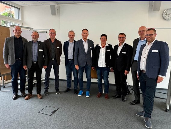 Neun Personen stehen in einem Besprechungsraum vor einer weißen Wand mit Flipcharts und Pinnwänden. Sie tragen Namensschilder und Business-Kleidung. Einige tragen Blazer, andere sportliche Jacken. Eine Uhr an der Wand zeigt die Zeit. Die Gruppe lächelt in die Kamera.