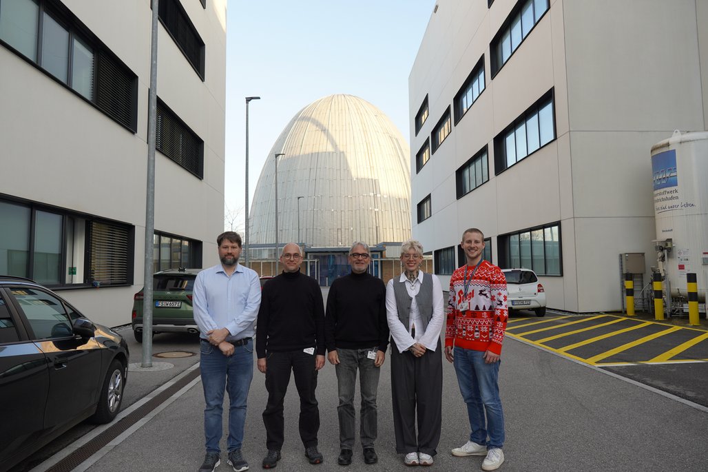Fünf Personen posieren vor modernen Institutsgebäuden und der markanten Atom-Ei-Kuppel in Garching.