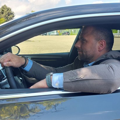 Ein Mann mit kurzem Bart sitzt in einem schwarzen Auto mit geöffnetem Fenster. Er trägt ein hellblaues Hemd mit einem braunen Sakko und eine Smartwatch am Handgelenk. Er lehnt entspannt mit einem Arm aus dem Fenster und hat die andere Hand am Lenkrad, während er mit einem konzentrierten Blick nach vorne schaut. Im Hintergrund ist ein Parkplatz mit mehreren Autos und einem mehrstöckigen Parkhaus zu sehen. Der Himmel ist leicht bewölkt, und das Sonnenlicht spiegelt sich im Autolack.