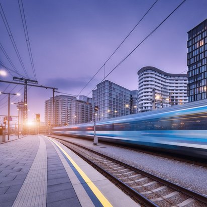 Ein moderner, blau-weißer Zug fährt mit hoher Geschwindigkeit durch einen städtischen Bahnhof bei Dämmerung. Die Bewegung des Zuges ist als Unschärfe sichtbar. Im Hintergrund stehen moderne Hochhäuser mit beleuchteten Fenstern. Der Himmel zeigt einen violett-blauen Farbton, während die untergehende Sonne links im Bild einen warmen Lichtschein erzeugt. Oberleitungen und Bahnsteigmarkierungen sind im Vordergrund sichtbar.