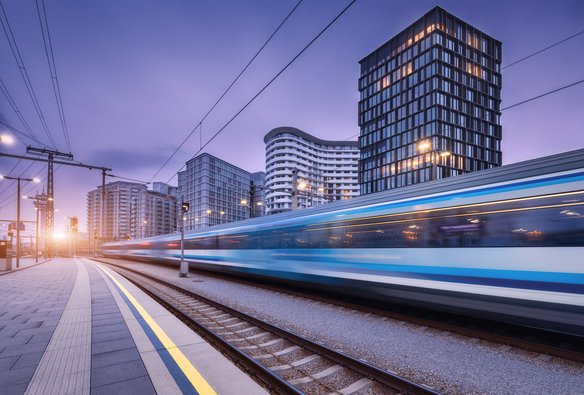 Ein moderner, blau-weißer Zug fährt mit hoher Geschwindigkeit durch einen städtischen Bahnhof bei Dämmerung. Die Bewegung des Zuges ist als Unschärfe sichtbar. Im Hintergrund stehen moderne Hochhäuser mit beleuchteten Fenstern. Der Himmel zeigt einen violett-blauen Farbton, während die untergehende Sonne links im Bild einen warmen Lichtschein erzeugt. Oberleitungen und Bahnsteigmarkierungen sind im Vordergrund sichtbar.