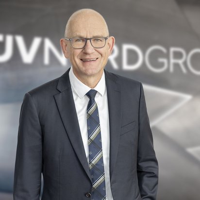 Ein lächelnder Mann mit Glatze und Brille trägt einen dunkelblauen Anzug, ein weißes Hemd und eine gemusterte Krawatte. Er steht vor einer modernen, metallischen Wand mit dem Schriftzug ‚TÜV NORD GROUP‘