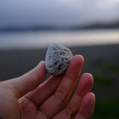 Eine Nahaufnahme einer Hand, die einen kleinen, grauen Stein mit sichtbaren Poren und Texturen hält. Der Stein ist im Bildmittelpunkt vor einem verschwommenen Hintergrund eines ruhigen Sees und entfernter Hügel. Das Licht ist weich und diffus und betont die Details des Steins."