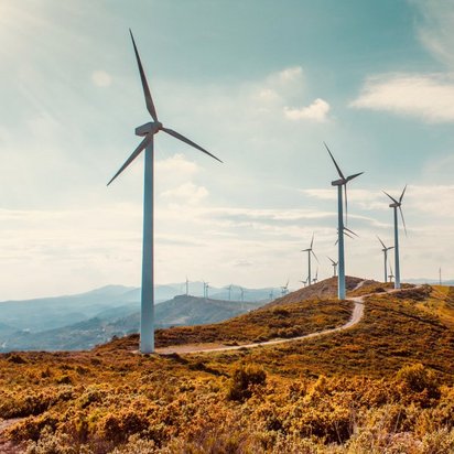Mehrere Windkraftanlagen stehen auf einer hügeligen Landschaft mit grün-brauner Vegetation. Ein schmaler Weg schlängelt sich durch die Hügel. Der Himmel ist teils bewölkt, und die Sonne wirft ein warmes Licht auf die Szenerie. Im Hintergrund erstrecken sich weitere Hügel und Windräder in der Ferne.