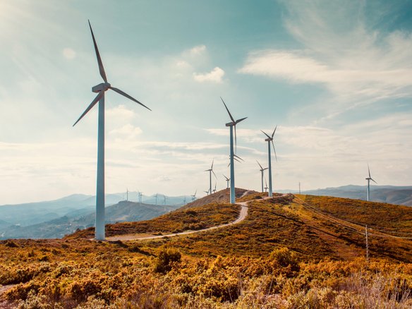 Mehrere Windkraftanlagen stehen auf einer hügeligen Landschaft mit grün-brauner Vegetation. Ein schmaler Weg schlängelt sich durch die Hügel. Der Himmel ist teils bewölkt, und die Sonne wirft ein warmes Licht auf die Szenerie. Im Hintergrund erstrecken sich weitere Hügel und Windräder in der Ferne.