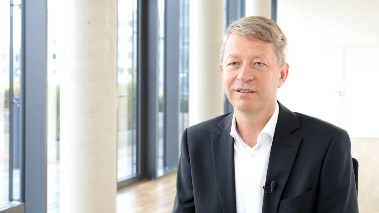Das Bild zeigt Ralf Dollenberg, International Business Development Manager, in einem modernen, lichtdurchfluteten Bürogebäude. Er trägt ein weißes Hemd und ein dunkles Sakko, auf dem Revers ist ein Ansteckmikrofon befestigt. Sein Blick ist offen und freundlich, während er leicht seitlich zur Kamera spricht. Im Hintergrund sind große Fensterfronten mit Betonpfeilern und Parkettboden zu sehen, die eine helle, professionelle Atmosphäre schaffen. Das Bild ist Teil eines Mitarbeiter-Testimonials und vermittelt Internationalität, Erfahrung und strategische Kompetenz.