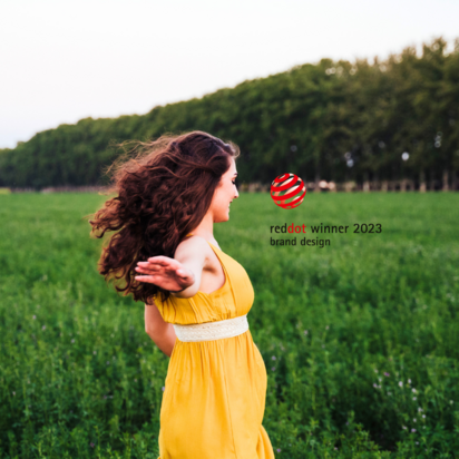Eine junge Frau mit lockigen braunen Haaren trägt ein gelbes Kleid und dreht sich mit ausgebreiteten Armen in einer grünen Wiese. Im Hintergrund sind Bäume und ein klarer Himmel zu sehen. Rechts im Bild befindet sich das Logo des Red Dot Design Awards mit der Aufschrift „reddot winner 2023 – brand design“.