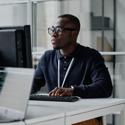 Ein junger Mann mit Brille und dunklem Polohemd sitzt an einem Schreibtisch in einem modernen Büro. Er arbeitet konzentriert an einem Computer mit zwei Bildschirmen, auf denen Programmiercode zu sehen ist. Vor ihm steht zudem ein aufgeklappter Laptop. Im Hintergrund befinden sich Glaswände und Regale mit Büromaterial. Die Szene wirkt professionell und technologisch geprägt.