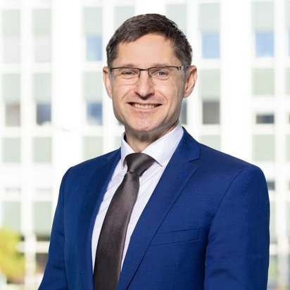 Porträt eines lächelnden Mannes mit kurzer dunkler Frisur und Brille, gekleidet in einen blauen Anzug mit weißem Hemd und dunkler Krawatte. Er steht vor einem großen Fenster mit Blick auf ein modernes Bürogebäude.