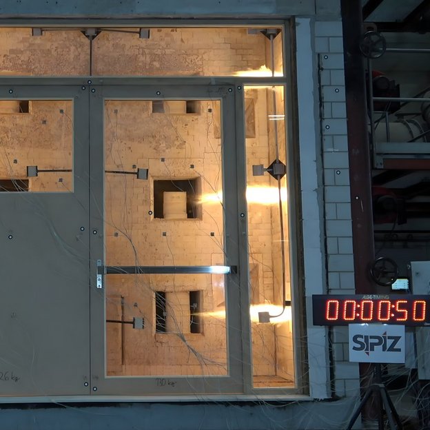 Eine große Tür- und Fensterkonstruktion wird in einem Labor einem Feuerwiderstandsversuch unterzogen. Dahinter leuchtet eine intensive Hitzequelle, während Sensoren und Kabel an der Tür befestigt sind. Rechts zeigt eine digitale Zeitanzeige ‚00:00:50‘ und das Logo ‚SPIZ‘ ist darunter zu sehen.