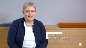 Das Bild zeigt Dr. med. Petra Jürgens, eine Ärztin für Verkehrsmedizin, die in einem neutral gestalteten Raum sitzt. Sie trägt eine Brille, eine dunkelblaue Strickjacke über einem weißen Oberteil sowie eine farblich abgestimmte Halskette. Im Hintergrund befinden sich ein helles Holzmöbel mit gepolsterten Sitzflächen in Orange und Blau sowie eine helle Wand. Petra Jürgens blickt freundlich in die Kamera. Der eingeblendete Text rechts im Bild lautet: ‚Dr. med. Petra Jürgens – medizinische Gutachterin‘. Das Thumbnail vermittelt Seriosität, Expertise und Empathie im Bereich der verkehrsmedizinischen Begutachtung.