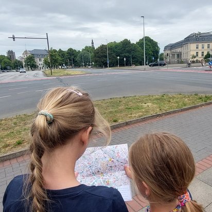 Zwei Kinder mit blonden Haaren stehen mit dem Rücken zur Kamera und halten gemeinsam eine Stadtkarte in den Händen. Sie stehen auf einem Bürgersteig vor einer großen Straßenkreuzung mit Fahrradwegen und Fußgängerüberwegen. Im Hintergrund sind Gebäude, Bäume und einige Menschen zu sehen.
