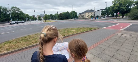 Zwei Kinder mit blonden Haaren stehen mit dem Rücken zur Kamera und halten gemeinsam eine Stadtkarte in den Händen. Sie stehen auf einem Bürgersteig vor einer großen Straßenkreuzung mit Fahrradwegen und Fußgängerüberwegen. Im Hintergrund sind Gebäude, Bäume und einige Menschen zu sehen.