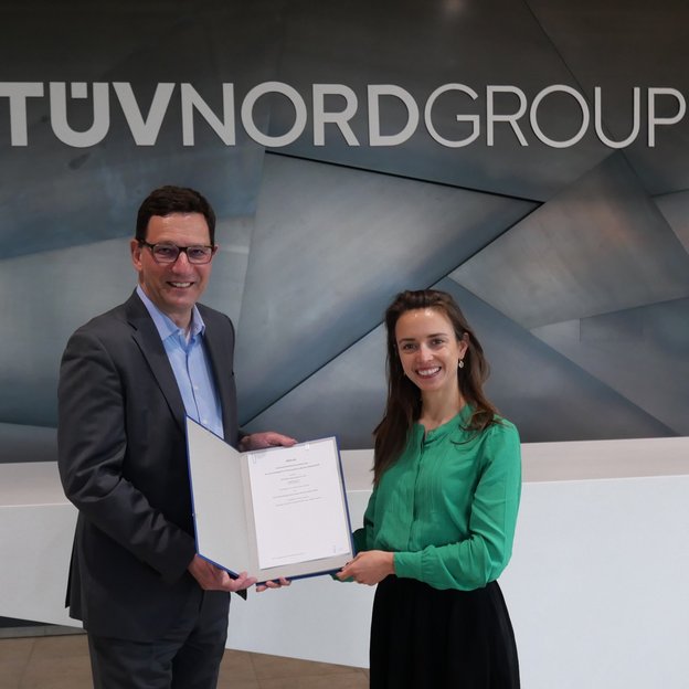 Ein Mann in einem grauen Anzug mit Brille und eine Frau in einer grünen Bluse stehen vor einer modernen Metallwand mit dem Schriftzug ‚TÜV NORD GROUP‘. Sie halten gemeinsam eine geöffnete Urkunde und lächeln in die Kamera.