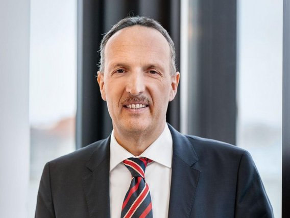 „Ein Mann mit kurzem, dunklem Haar und Oberlippenbart steht vor einem großen Fenster. Er trägt einen dunkelblauen Anzug mit weißem Hemd und eine rot-schwarz gestreifte Krawatte. Er blickt freundlich in die Kamera.