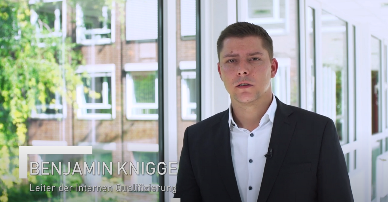 Das Bild zeigt Benjamin Knigge, Leiter der internen Qualifizierung, in einem hellen Flur mit großen Fensterfronten. Er trägt ein dunkles Sakko über einem weißen Hemd und blickt ruhig in Richtung Kamera. Ein Ansteckmikrofon ist am Revers befestigt. Durch die Fenster ist ein Gebäude mit roten Klinkerwänden und weißen Fenstern sichtbar, umrahmt von grüner Vegetation. Die Lichtstimmung ist freundlich und professionell. Das Thumbnail gehört zu einem Mitarbeiter-Testimonial, das Einblicke in interne Weiterbildungs- und Qualifizierungsprozesse gibt.