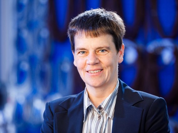 Eine lächelnde Frau mit kurzen, dunklen Haaren trägt einen dunkelblauen Blazer über einer gestreiften Bluse. Sie steht vor einem unscharfen technischen Hintergrund mit blauen und schwarzen Elementen.“