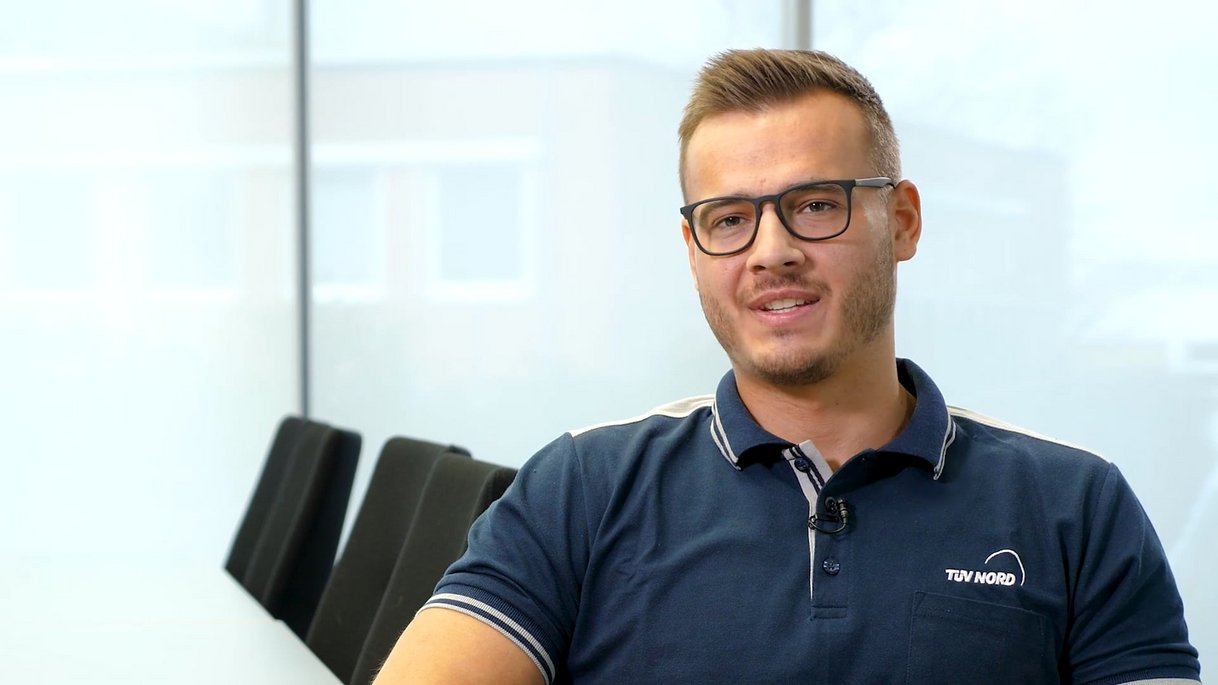 Das Bild zeigt einen jungen Mann mit kurzen, blond-braunen Haaren und Brille, der freundlich in die Kamera blickt. Er trägt ein dunkelblaues Polo-Shirt mit dem TÜV NORD Logo auf der linken Brustseite. Der Hintergrund ist ein modernes, lichtdurchflutetes Büro mit Glaswänden und schlichten schwarzen Stühlen. Dieses Thumbnail gehört zu einem Testimonial eines Auszubildenden zum Prüfingenieur und vermittelt eine professionelle, aber zugleich angenehme Arbeitsatmosphäre.