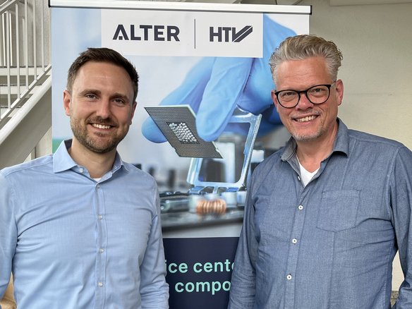 Zwei Männer stehen lächelnd vor einem Banner mit den Logos von ‚ALTER‘ und ‚HTV‘. Der Mann links trägt ein hellblaues Hemd, der Mann rechts ein dunkelblaues Hemd und eine Brille. Im Hintergrund zeigt das Banner ein technisches Bild mit einem Mikrochip und einer Hand in einem blauen Handschuh