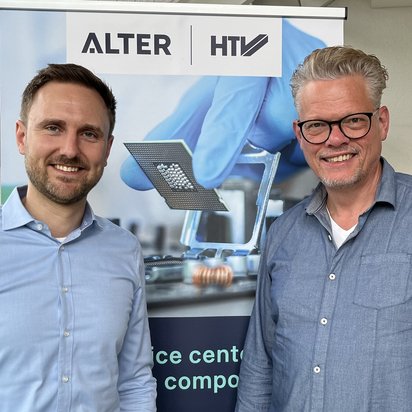 Zwei Männer stehen lächelnd vor einem Banner mit den Logos von ‚ALTER‘ und ‚HTV‘. Der Mann links trägt ein hellblaues Hemd, der Mann rechts ein dunkelblaues Hemd und eine Brille. Im Hintergrund zeigt das Banner ein technisches Bild mit einem Mikrochip und einer Hand in einem blauen Handschuh