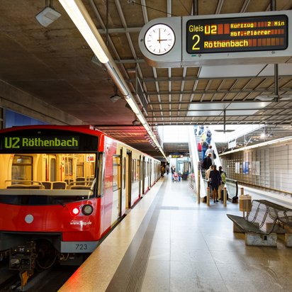 Eine rote U-Bahn der Linie U2 mit der Zielanzeige „Röthenbach“ steht an einem unterirdischen Bahnsteig. Die Station hat eine moderne Architektur mit blauen Wandfliesen und einer Rolltreppe im Hintergrund. Menschen steigen ein und aus, einige benutzen die Rolltreppe. Eine elektronische Anzeige zeigt Abfahrtsinformationen an.