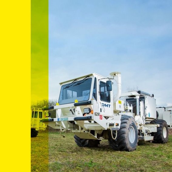 A white industrial DMT vehicle with large tires and a boxy cabin is parked on a grassy field, 