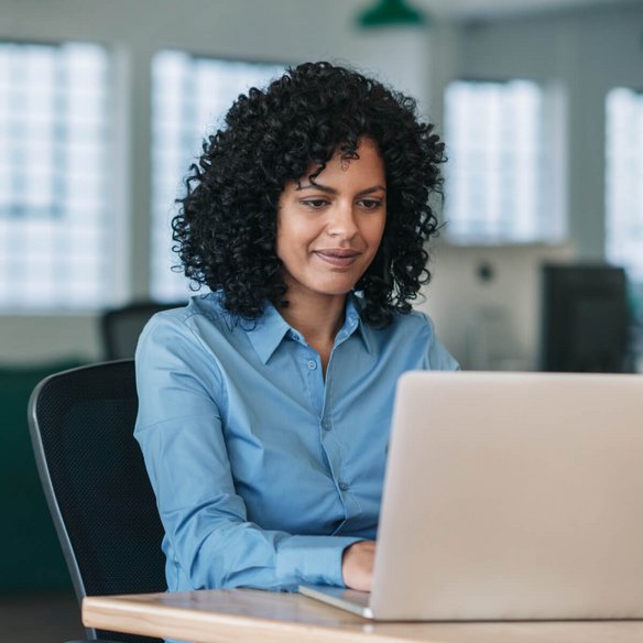 Eine Frau mit lockigen dunklen Haaren sitzt an einem Schreibtisch in einem modernen Büro. Sie trägt eine hellblaue Bluse und arbeitet konzentriert an einem silbernen Laptop. Ihr Gesichtsausdruck ist freundlich und fokussiert. Im Hintergrund sind große Fenster, mehrere Schreibtische und Büroeinrichtungen zu sehen, die eine offene und helle Arbeitsatmosphäre schaffen.