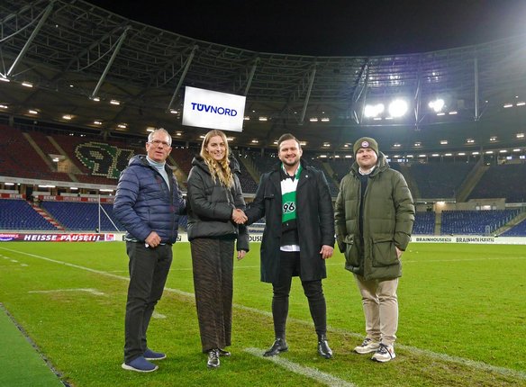 Vier Personen stehen auf dem Rasen des Stadions von Hannover 96 und schütteln sich die Hände. Im Hintergrund sind die leeren Tribünen und eine große Anzeigetafel mit dem TÜV NORD-Logo zu sehen. Die Personen tragen warme Kleidung, darunter eine Hannover 96-Fanschal. Das Stadion ist beleuchtet, und die Atmosphäre deutet auf eine offizielle Partnerschaft oder ein Sponsoring hin.