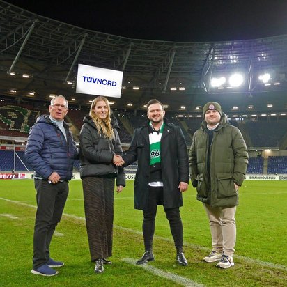 Vier Personen stehen auf dem Rasen des Stadions von Hannover 96 und schütteln sich die Hände. Im Hintergrund sind die leeren Tribünen und eine große Anzeigetafel mit dem TÜV NORD-Logo zu sehen. Die Personen tragen warme Kleidung, darunter eine Hannover 96-Fanschal. Das Stadion ist beleuchtet, und die Atmosphäre deutet auf eine offizielle Partnerschaft oder ein Sponsoring hin.