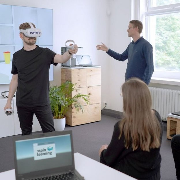 Ein Mann trägt eine VR-Brille mit dem Logo „TÜV NORD“ und hält zwei VR-Controller in den Händen, während er eine virtuelle Umgebung erkundet. Auf einem großen Bildschirm im Hintergrund wird die VR-Simulation angezeigt. Drei Personen beobachten ihn aufmerksam – eine Frau mit langen Haaren und zwei Männer, einer davon spricht und gestikuliert. Im Vordergrund steht ein Laptop mit dem Logo „3spin Learning“. Der Raum ist modern eingerichtet mit weißen Wänden, großen Fenstern und Holzmöbeln.