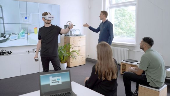 Ein Mann trägt eine VR-Brille mit dem Logo „TÜV NORD“ und hält zwei VR-Controller in den Händen, während er eine virtuelle Umgebung erkundet. Auf einem großen Bildschirm im Hintergrund wird die VR-Simulation angezeigt. Drei Personen beobachten ihn aufmerksam – eine Frau mit langen Haaren und zwei Männer, einer davon spricht und gestikuliert. Im Vordergrund steht ein Laptop mit dem Logo „3spin Learning“. Der Raum ist modern eingerichtet mit weißen Wänden, großen Fenstern und Holzmöbeln.
