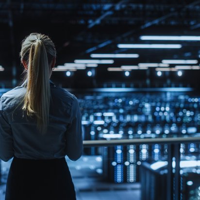 Eine Frau mit langen, blond gebundenen Haaren steht mit dem Rücken zur Kamera und blickt auf ein großes, modern beleuchtetes Rechenzentrum mit zahlreichen Serverracks und leuchtenden LED-Anzeigen.