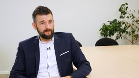 Das Bild zeigt Pierre Fontana, Senior Test Engineer, in einem modernen Besprechungsraum. Er trägt ein dunkelblaues Sakko über einem weißen Hemd und sitzt entspannt an einem großen Konferenztisch. Er hat dunkles, kurz geschnittenes Haar und einen gepflegten Bart. Im Hintergrund sind schwarze Stühle, eine helle Wand und eine grüne Zimmerpflanze zu sehen. Die Stimmung des Bildes ist ruhig und professionell. Das Bild ist Teil eines Mitarbeiter-Testimonials und vermittelt Fachkompetenz und Vertrauen.