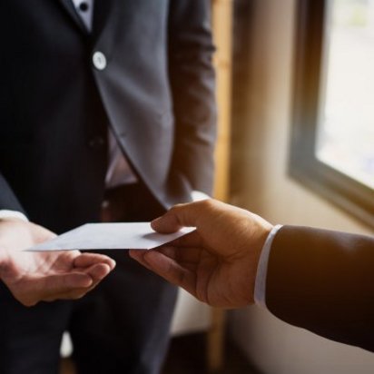 Zwei Personen in dunklen Anzügen stehen sich gegenüber. Eine Hand hält einen weißen Umschlag und übergibt ihn der anderen Person, die die Hand geöffnet hält, um ihn anzunehmen. Die Szene wirkt diskret und findet in einem Raum mit Fensterlicht statt.