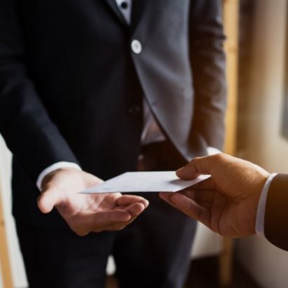 Zwei Personen in dunklen Anzügen stehen sich gegenüber. Eine Hand hält einen weißen Umschlag und übergibt ihn der anderen Person, die die Hand geöffnet hält, um ihn anzunehmen. Die Szene wirkt diskret und findet in einem Raum mit Fensterlicht statt.