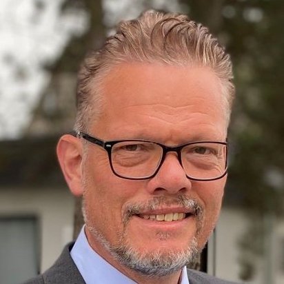 Porträt eines Mannes mit kurzen, nach hinten gestylten Haaren, Brille und grauem Bart. Er trägt einen dunklen Anzug mit einem hellblauen Hemd und eine gemusterte Krawatte. Im Hintergrund ist ein Gebäude mit Fenstern und Bäumen leicht unscharf zu sehen