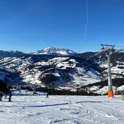 Winterliche Berglandschaft mit schneebedeckten Bergen und einem strahlend blauen Himmel. Im Vordergrund befinden sich mehrere Skifahrer auf einer Piste. Rechts im Bild verläuft eine Gondelbahn mit einer großen Stütze, die eine Seilbahn trägt. Im Hintergrund erstreckt sich ein Tal mit verschneiten Hügeln und Wäldern, während die Sonne die Alpenlandschaft erhellt.