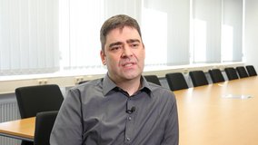 Das Bild zeigt Stefan Slabihoud, Projektleiter für Softwareprodukte, Leiter des Softwarelabors und Senior Consultant bei der TÜV NORD GROUP. Er sitzt in einem modernen, hellen Konferenzraum mit langen Tischen und schwarzen Stühlen. Die Fenster im Hintergrund sind mit vertikalen Lamellen-Jalousien versehen. Stefan trägt ein dunkles Hemd und spricht in die Kamera, während er ernst und fokussiert wirkt. Das Bild ist Teil eines Mitarbeiter-Testimonials.