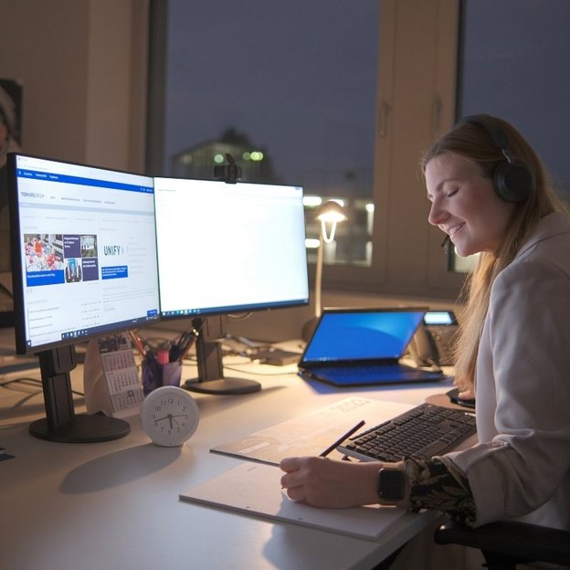 Eine junge Frau mit langen, offenen Haaren sitzt abends in einem Büro vor zwei Computerbildschirmen. Sie trägt ein Headset und lächelt, während sie Notizen auf einem Block macht. Auf ihrem Schreibtisch stehen ein Laptop, eine Lampe, ein Kalender, Stifte und eine kleine Uhr, die etwa 18:50 Uhr anzeigt. Im Hintergrund ist ein Fenster mit Blick auf eine beleuchtete Stadtlandschaft sowie ein TÜV NORD-Poster mit zwei Personen zu sehen.