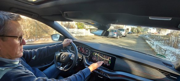 Ein Mann mit Brille und dunkelblauer Jacke fährt ein modernes Auto und bedient das Navigationssystem über den Touchscreen. Das Display zeigt eine Straßenkarte mit Routenführung. Durch die Windschutzscheibe ist eine Wohngegend mit parkenden Autos, Gehweg und leicht verschneitem Boden zu sehen.