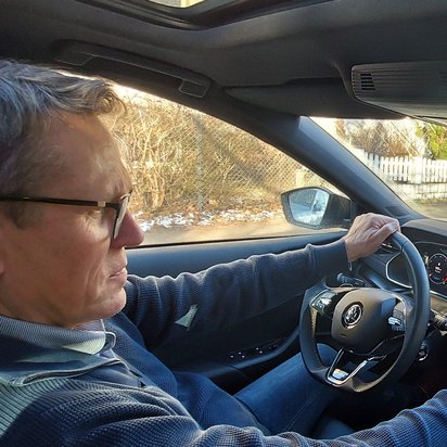 Ein Mann mit Brille und dunkelblauer Jacke fährt ein modernes Auto und bedient das Navigationssystem über den Touchscreen. Das Display zeigt eine Straßenkarte mit Routenführung. Durch die Windschutzscheibe ist eine Wohngegend mit parkenden Autos, Gehweg und leicht verschneitem Boden zu sehen.