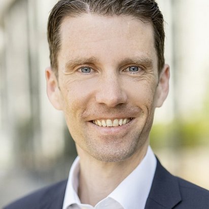 portraet-anatol-sendker-business Ein Mann mit kurzen, dunkelblonden Haaren lächelt freundlich in die Kamera. Er trägt ein dunkles Sakko über einem weißen Hemd. Der Hintergrund ist leicht unscharf und zeigt eine moderne Glasfassade mit Spiegelungen sowie grüne Elemente, die auf eine städtische Umgebung mit Bepflanzung hinweisen. Das Bild wirkt professionell und lichtdurchflutet.