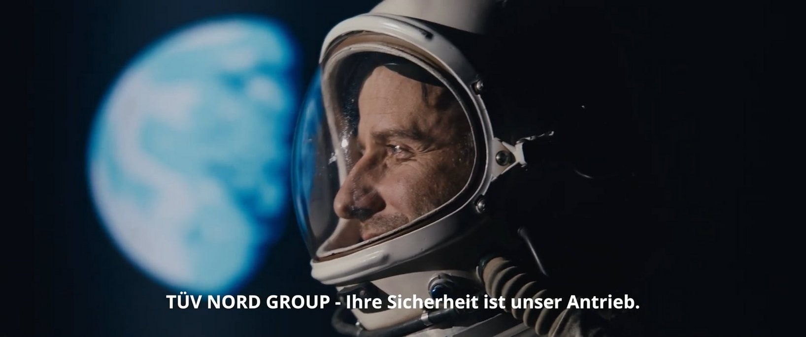 The picture shows a close-up of an astronaut with a white space helmet and reflective visor. He looks into the distance with a satisfied and thoughtful expression on his face, while the Earth can be seen blurred in the background. The scene is kept in dark colors, which enhances the contrast between the astronaut and the planet. The words ‘TÜV NORD GROUP - Your safety is our motivation’ are displayed at the bottom of the image. This image is the thumbnail for the TÜV NORD GROUP image film and symbolizes progress, innovation and safety.