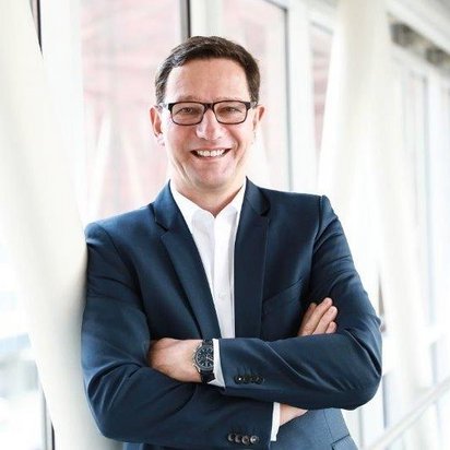 Ein Mann mit dunklen Haaren, Brille und einem dunkelblauen Anzug steht in einem modernen, lichtdurchfluteten Gebäude mit großen Fenstern. Er hat die Arme verschränkt und lächelt freundlich in die Kamera. Im Hintergrund sind Glasfassaden und architektonische Strukturen zu sehen.