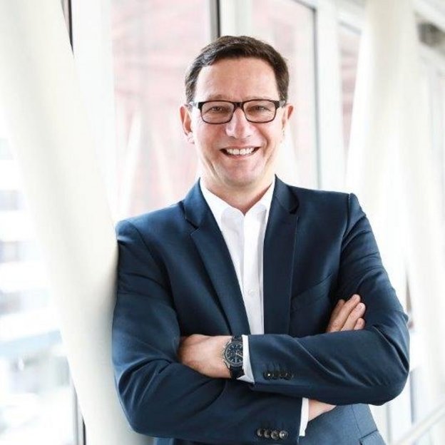 Ein Mann mit dunklen Haaren, Brille und einem dunkelblauen Anzug steht in einem modernen, lichtdurchfluteten Gebäude mit großen Fenstern. Er hat die Arme verschränkt und lächelt freundlich in die Kamera. Im Hintergrund sind Glasfassaden und architektonische Strukturen zu sehen.
