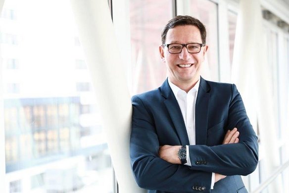 Ein Mann mit dunklen Haaren, Brille und einem dunkelblauen Anzug steht in einem modernen, lichtdurchfluteten Gebäude mit großen Fenstern. Er hat die Arme verschränkt und lächelt freundlich in die Kamera. Im Hintergrund sind Glasfassaden und architektonische Strukturen zu sehen.