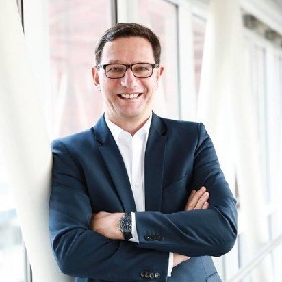 Ein Mann mit dunklen Haaren, Brille und einem dunkelblauen Anzug steht in einem modernen, lichtdurchfluteten Gebäude mit großen Fenstern. Er hat die Arme verschränkt und lächelt freundlich in die Kamera. Im Hintergrund sind Glasfassaden und architektonische Strukturen zu sehen.